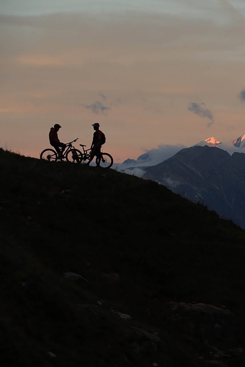 Two Santa Cruz mountain bikers standing on a hill, overlooking a mountain backdrop
