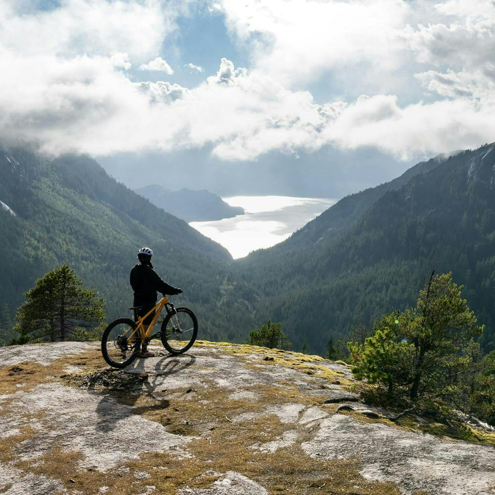 A mountain biker standing next to her bike looking out on aspen covered mountains and a lake