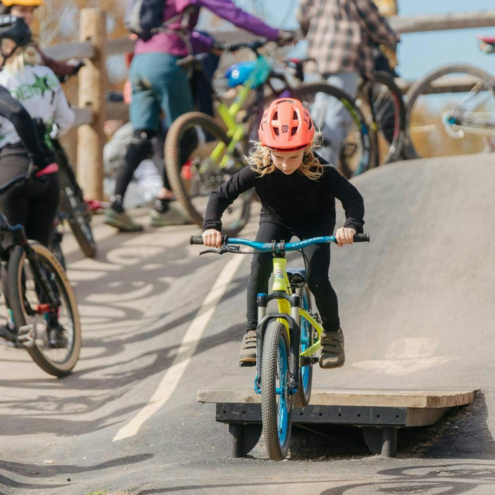 A kid riding her green bike off of a small wood drop on a paved pumptrack