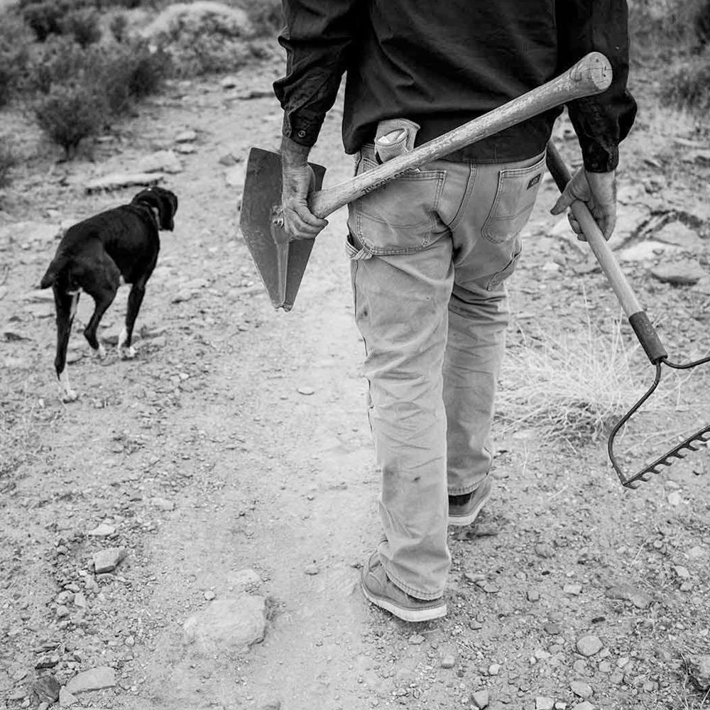 a trail builder holding tools and walking with their dog 