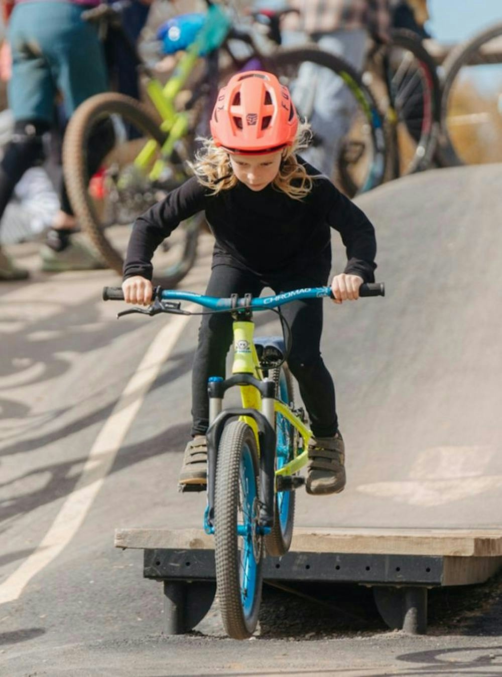 A young girl riding her bike off of a wood ramp on a paved pump track