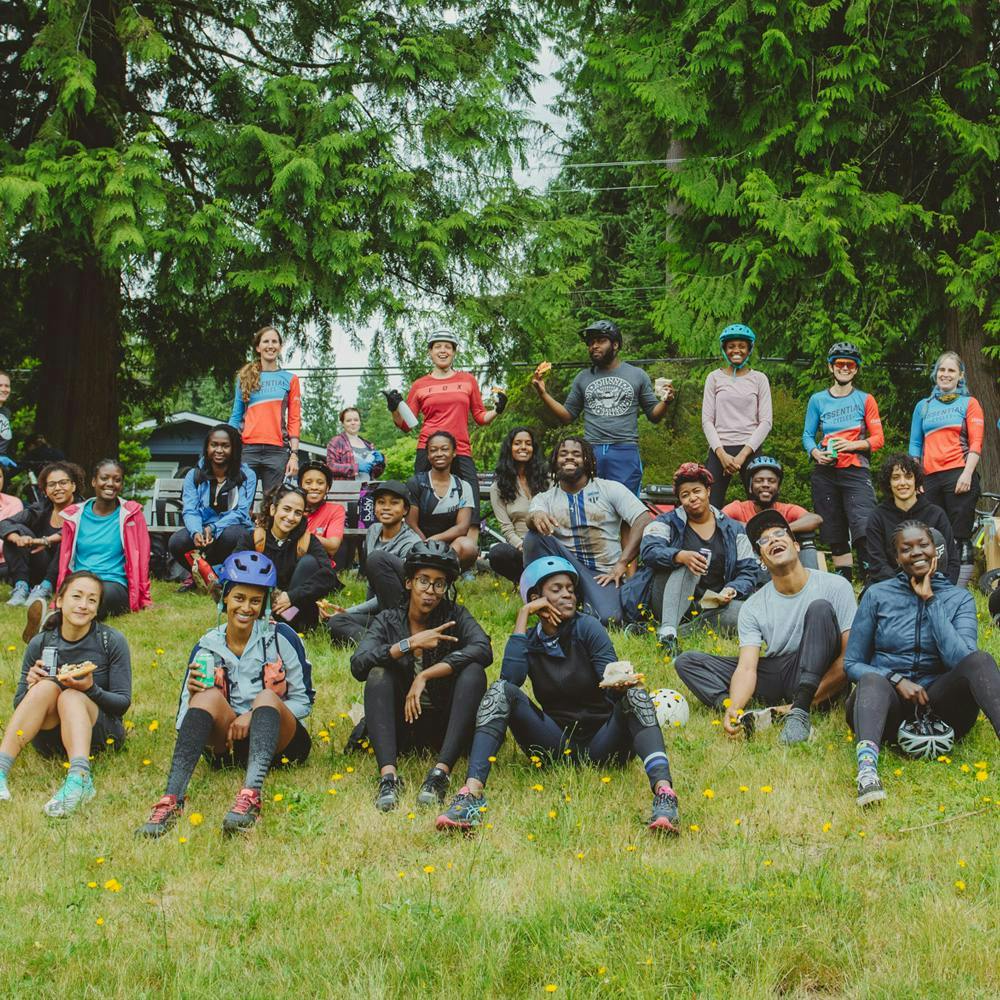 A group of BIPOC mountain bikers sitting on a grassy field, with a few mountain bikers standing behind them