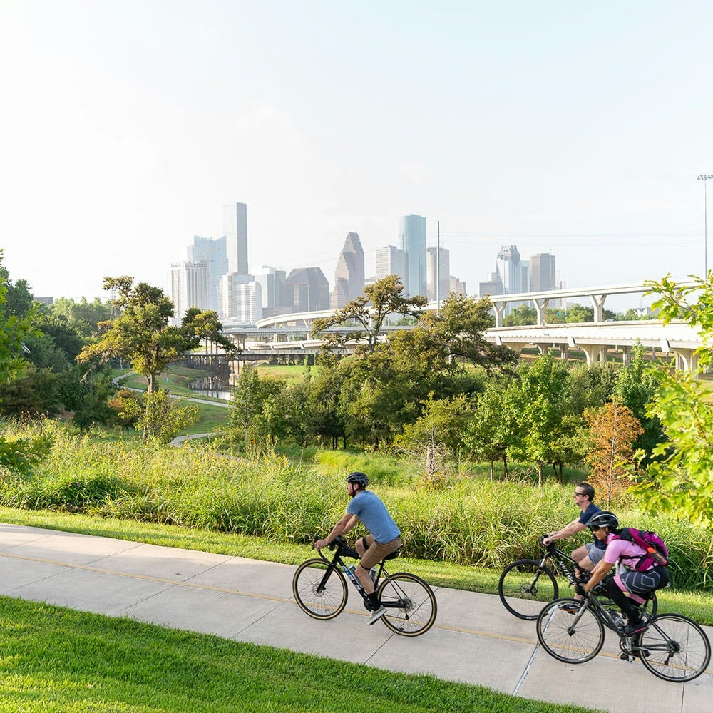 3 cyclists riding on a paved bike path with skyscrapers in the background