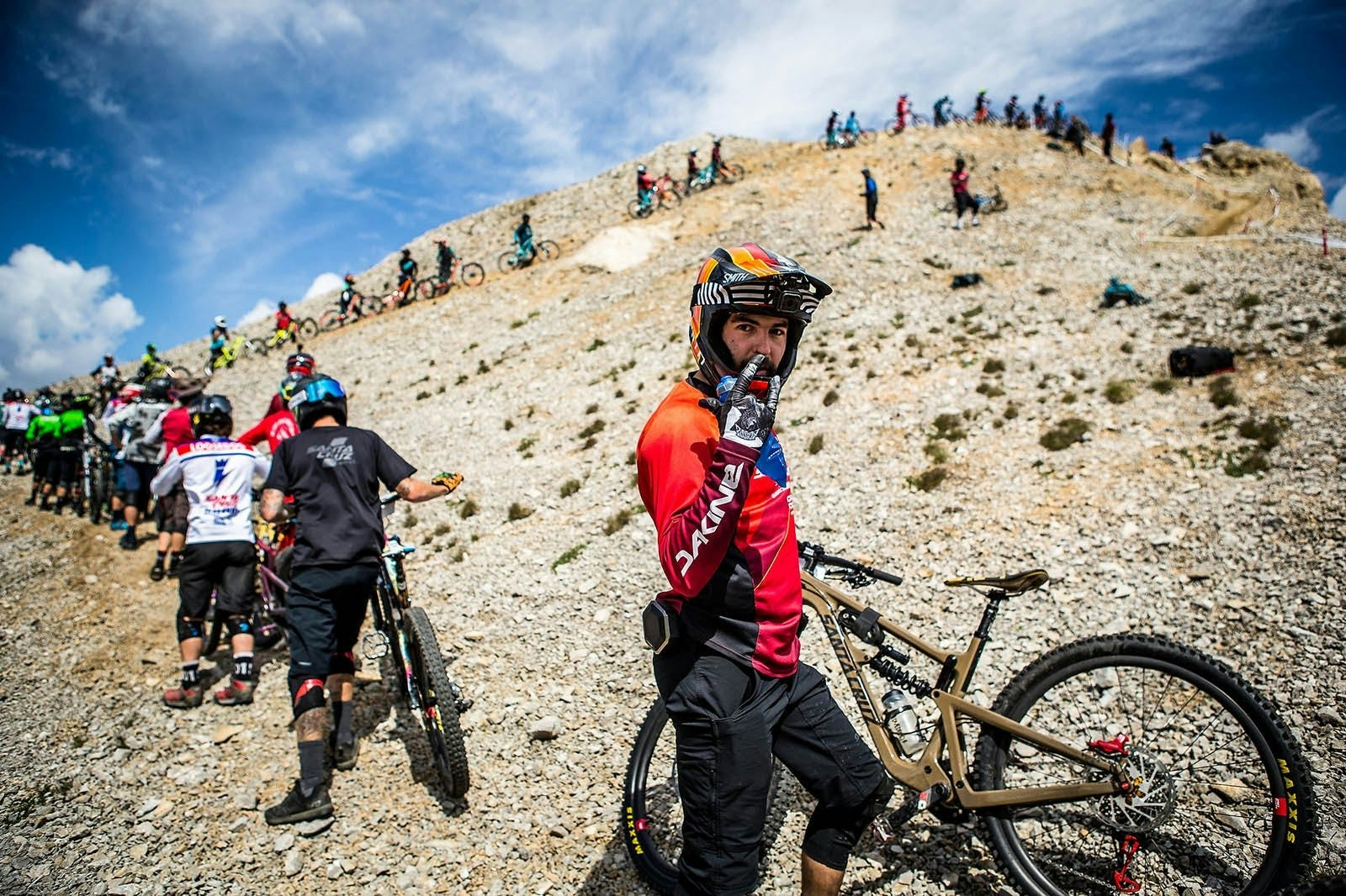 one mountain biker posing for the camera while many other hike their bikes up a hill