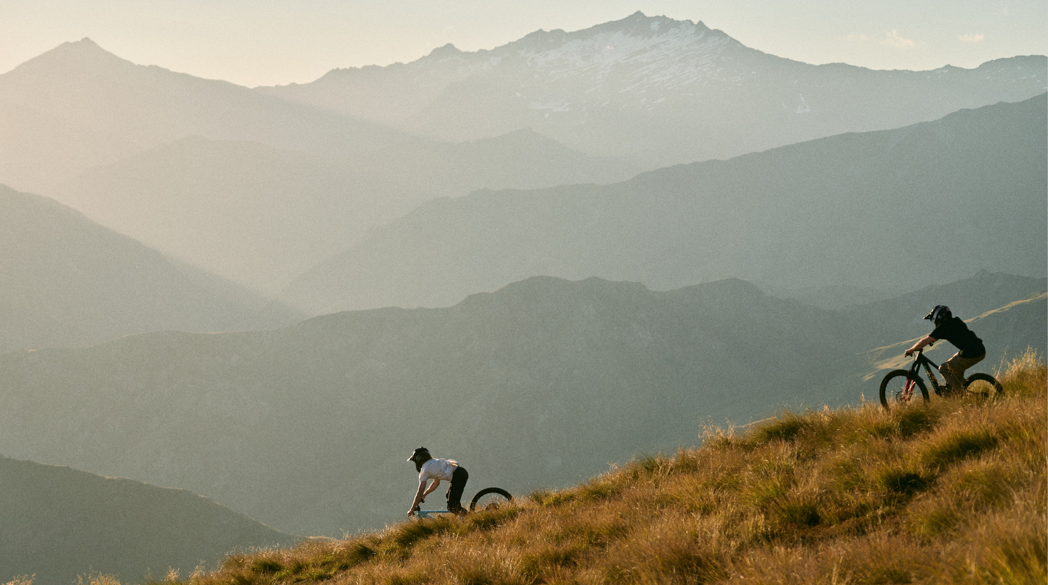 Two people with bicycles on a grassy hill with mountains in the background